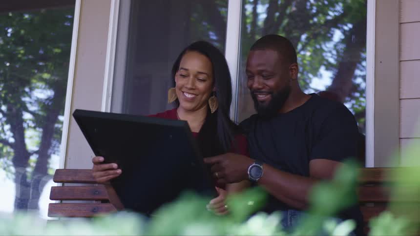 A man and a woman sitting on a bench in front of a house looking at a picture in a frame