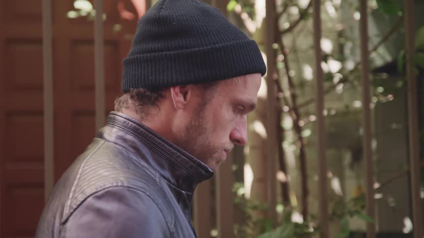 Profile of a man wearing a leather jacket and a beanie standing outside a house