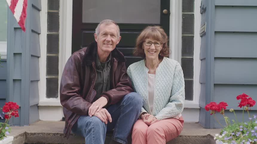 Happy elderly couple sitting on the steps at the entrance of a house smile looking at the camera