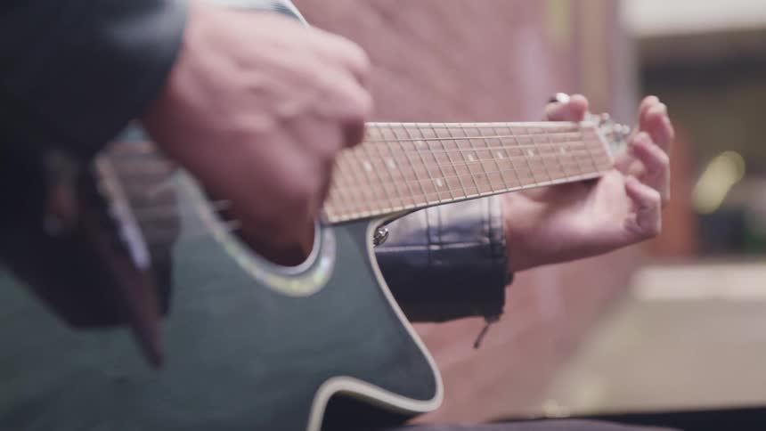 A person with a jacket playing an acoustic guitar next to a wall with a vehicle in the background