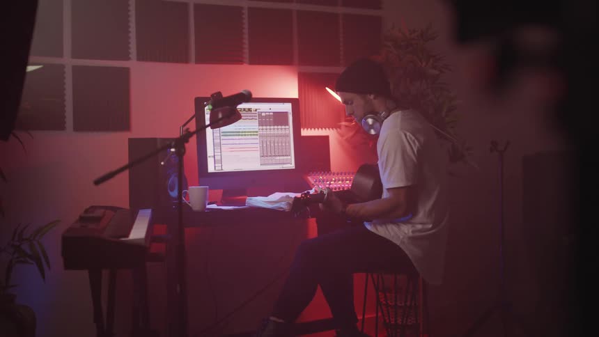 Young man sitting in front of a computer writes music while playing acoustic guitar in a studio