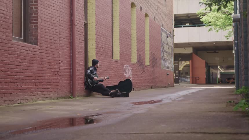 A man with a hat and a jacket sitting in an alleyway playing a guitar