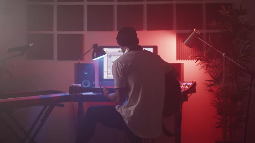 A man with a hat playing a guitar in a music studio in front of a computer with speakers