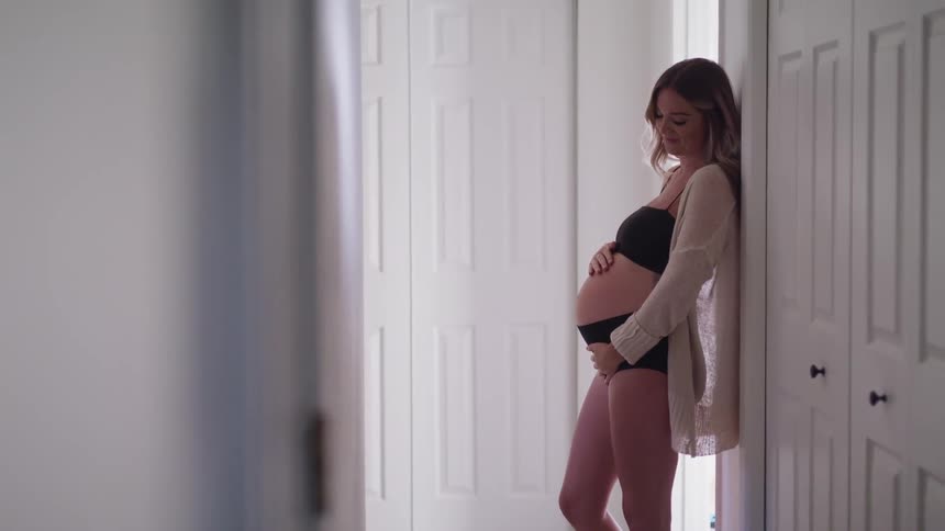 A pregnant woman rubbing her tummy while standing in a hallway in a bra and underwear