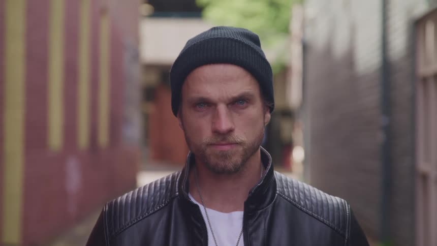 A man with a jacket and hat standing and staring in an alleyway