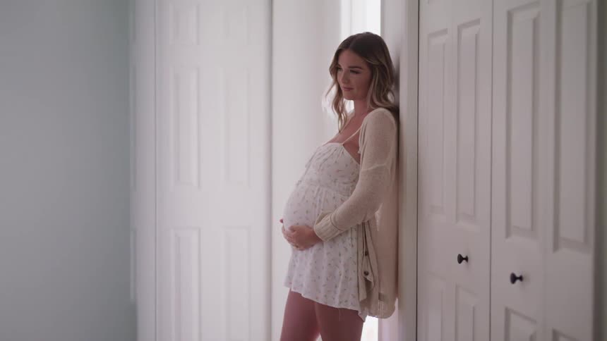 A woman at home leans against a wall and rubs her pregnant belly
