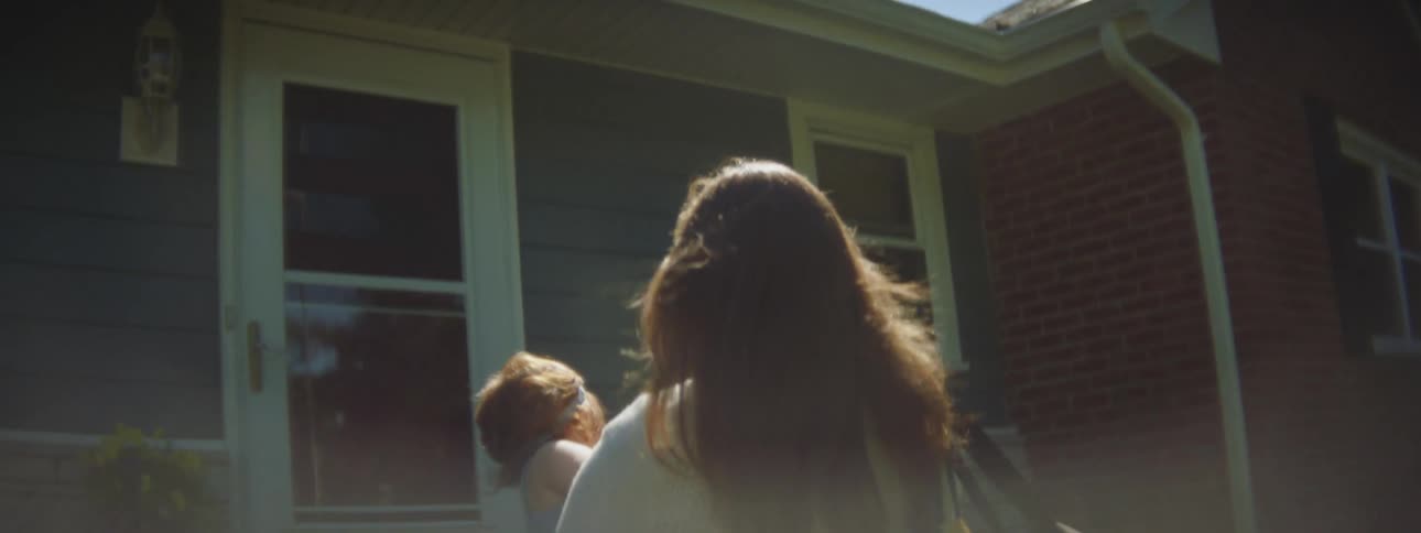 A mother walks towards a house and carries her infant daughter up the stairs to the front door