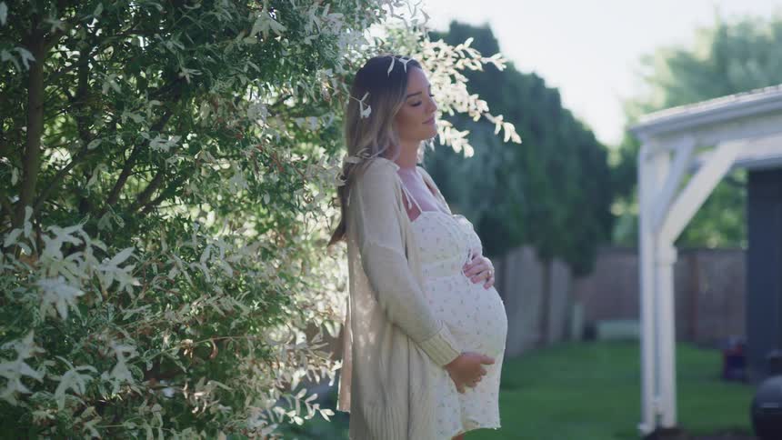 Young pregnant woman in a dress standing next to the white flowers on a tree in the backyard at evening