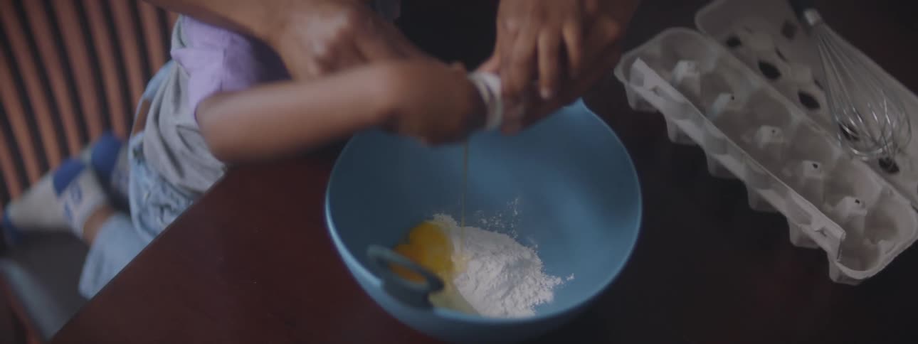 A mother helping a young child crack an egg into a bowl of flour