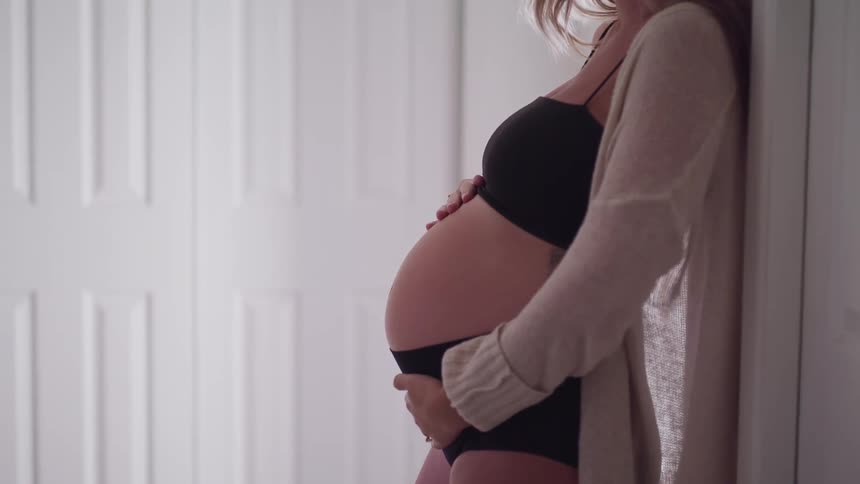 A pregnant woman leans against a wall at home and rubs her belly