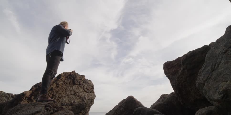 A man stands on a large rock taking a picture with his camera