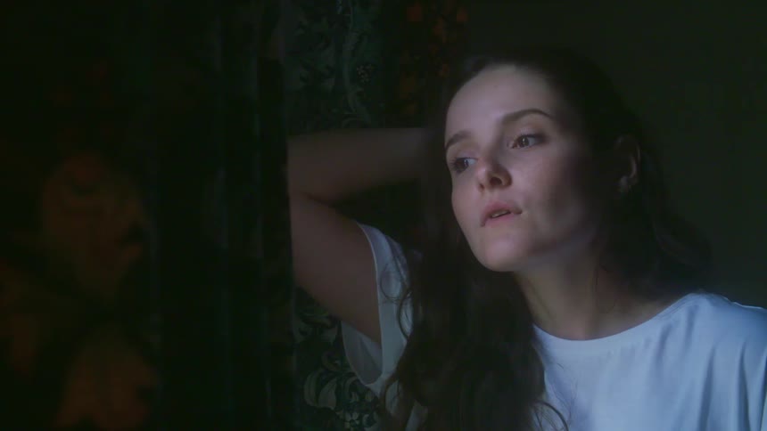 A young woman looking out a window in a dark room with soft light.
