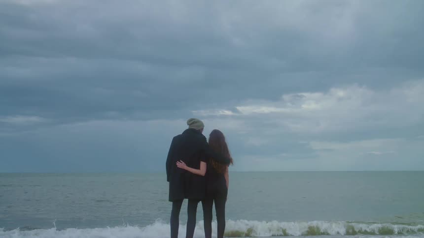 Man wearing beanie and black coat and woman with shirt and black pants holding each other watch the sea on the beach on a cloudy day