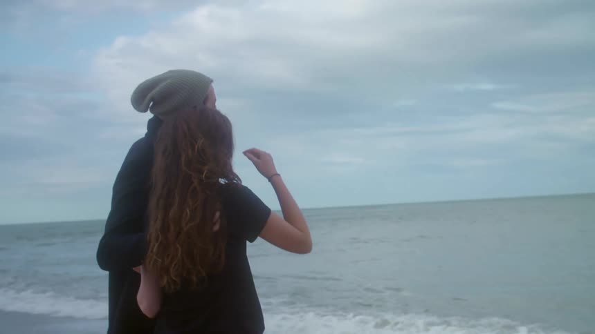 A man and a woman romantically hold one another while looking out at the ocean on a cloudy day