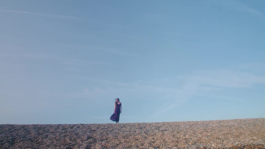 A woman in a long dress walks outdoors on a pebble beach on a windy day across an empty landscape