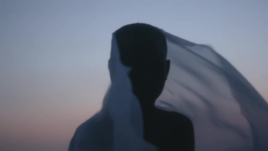 A boy standing by a shore with a sheet on him blowing in the wind with the sky and clouds in the background