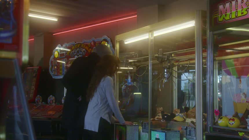 A man walks up to and hugs his girlfriend as she plays a claw machine in an arcade on a date