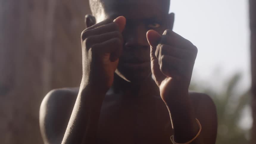 A boy throws punches at the air and holds a fighting stance while standing outdoors near a ruined building during the day