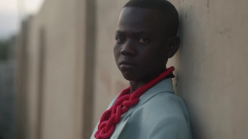A boy with a shirt and a rope around his neck standing by a wall with the sky and clouds in the background