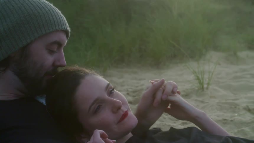 A man with a hat and a woman romantically laying on the sand by the beach during sunset.
