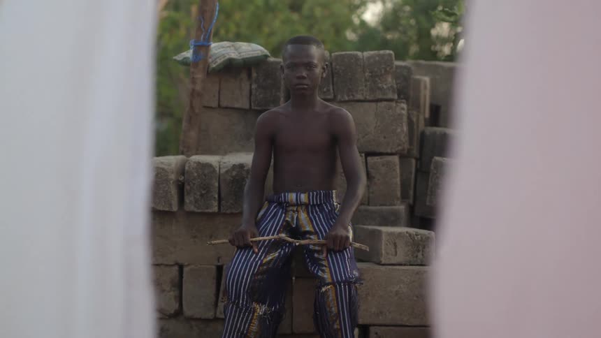 A young boy in an impoverished area sits on a pile of bricks without a shirt