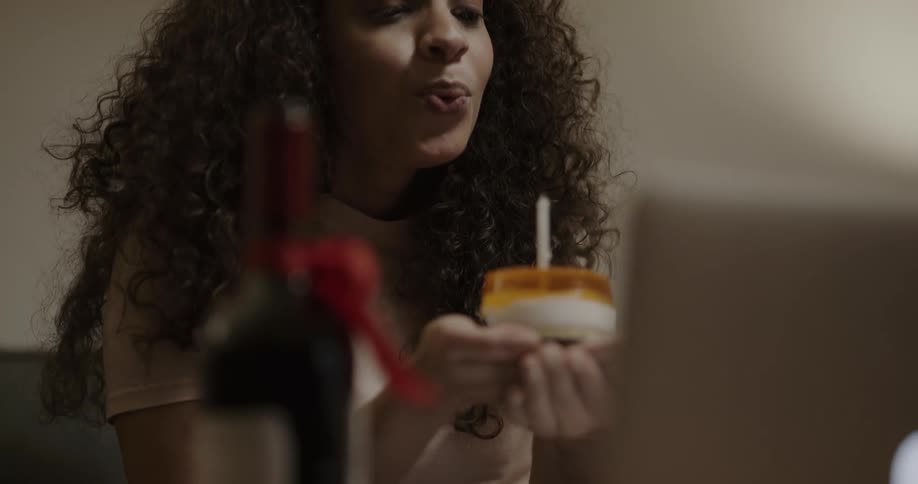 A woman in a room with a cake with a lit candle video chatting on a laptop