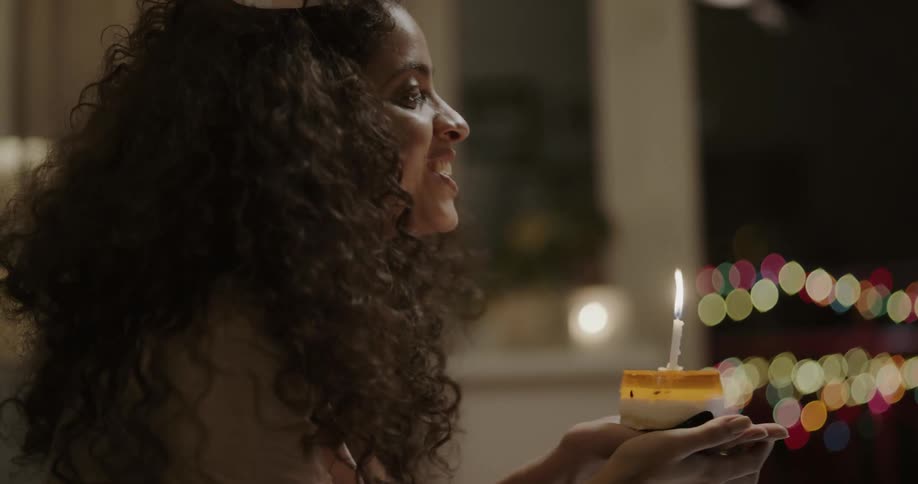 A woman in a room video chatting on a laptop holding a cake with a lit candle on it