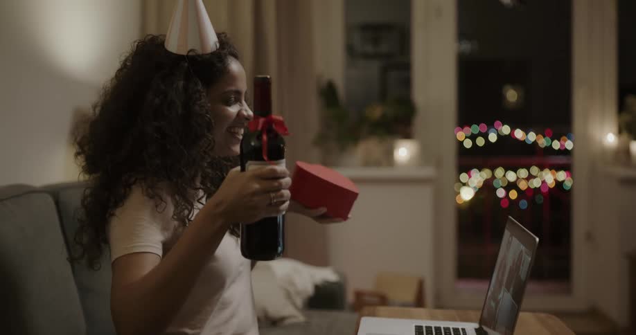 A young woman sitting on her couch celebrating with four friends on the computer with wine and a heart shaped box