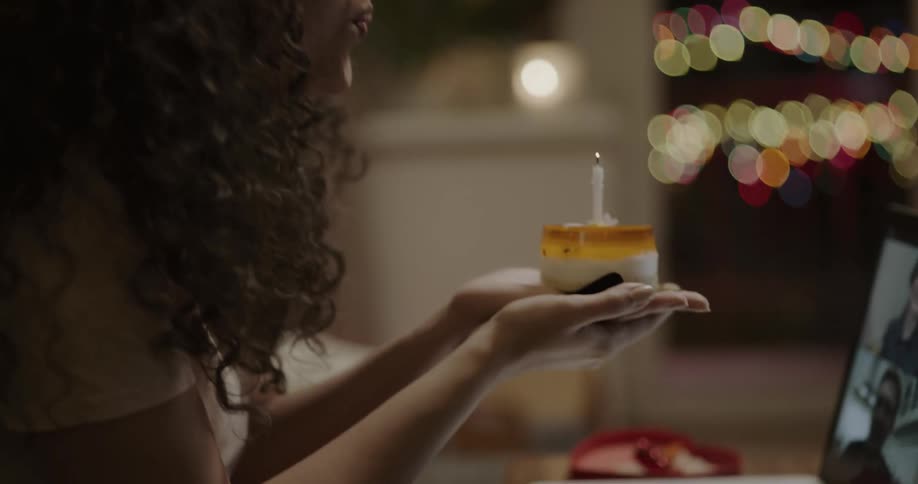 Young woman blowing the candle over a small pastry celebrating her birthday through a video call on a laptop
