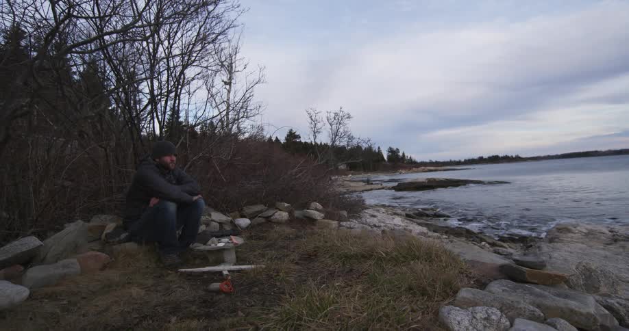 A man sits on a rock near a lake shore and looks forwards