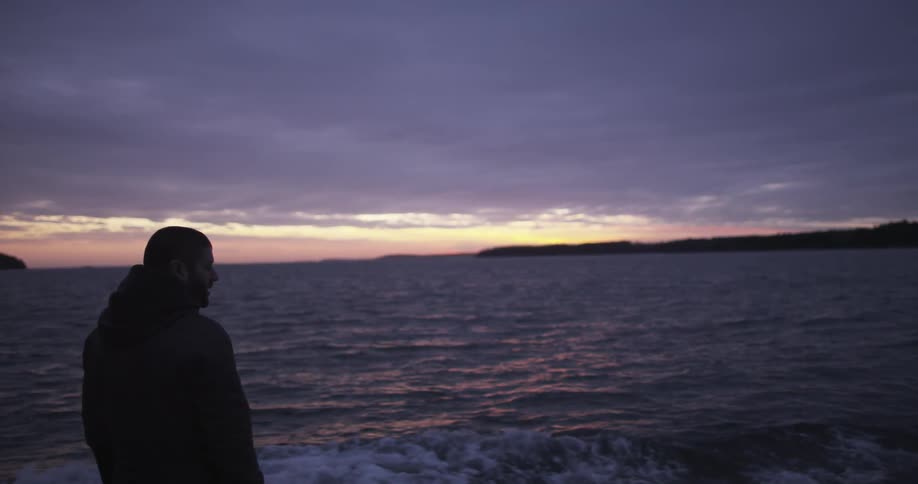 A man standing near the rail of a boat watching the waves at dusk