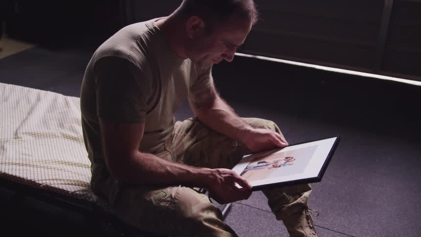 A man in a room with a military uniform sitting on a bed looking at a picture in a frame