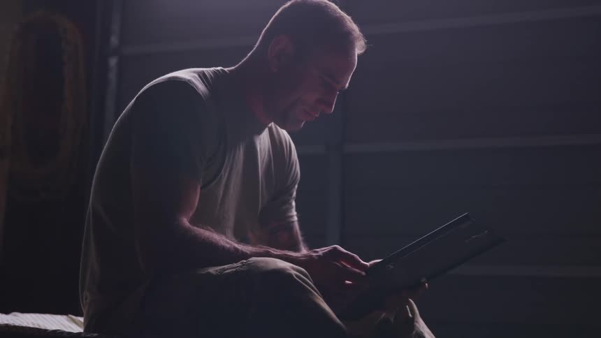 Soldier sitting in a bed reading the screen of a laptop in a dimly lit room