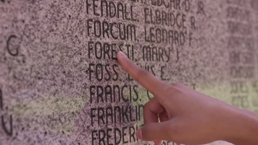 A woman runs her finger down a list of names on a monument
