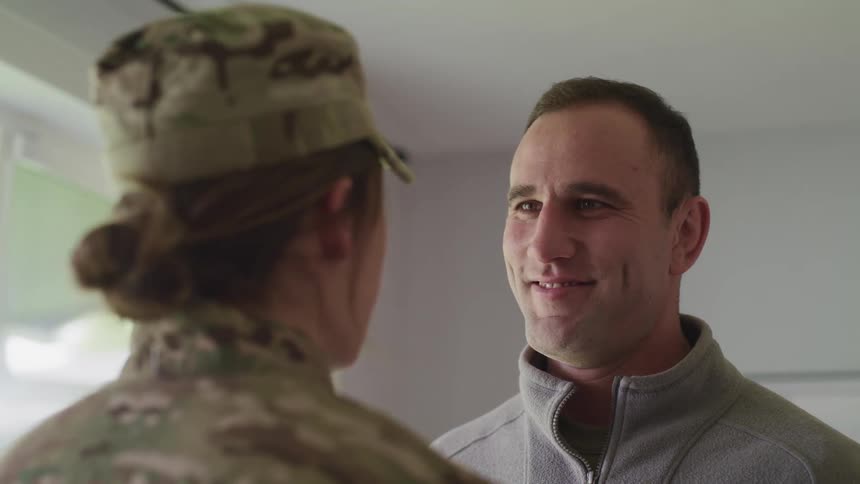 A man in a room standing and smiling at a woman in a military uniform with a window in the background
