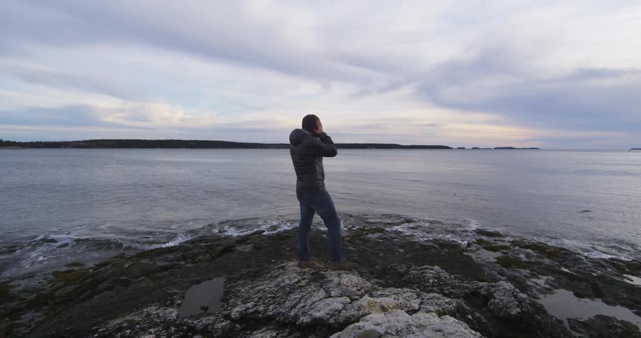 A man wearing a coat puts on a hat while standing near the shore