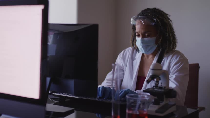 A woman in a lab coat and gloves sitting at a desk typing on a computer
