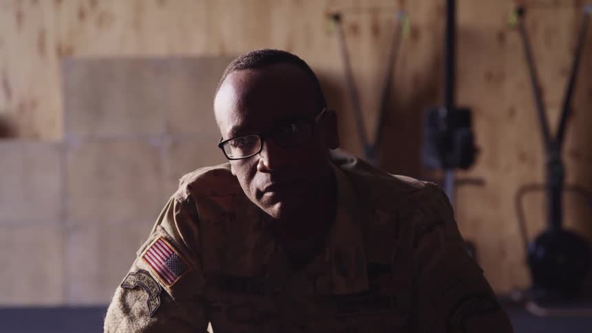 A soldier raises her hand to gaze into the camera while sitting indoors