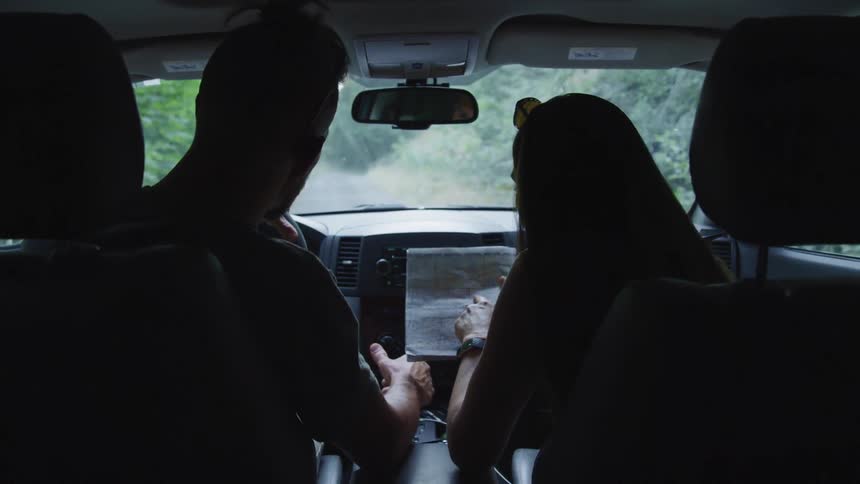 A man and a woman in a vehicle looking at a map while driving on a road surrounded by trees