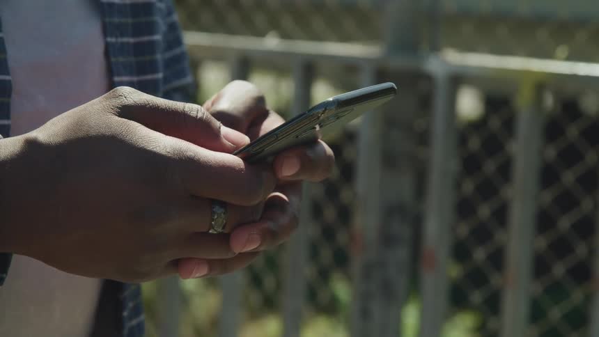 A person texting on a smartphone outside next to a chain link fence