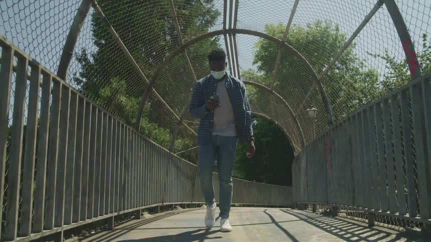 A man wearing a face mask looks down at his phone while walking over a gated bridge during the day
