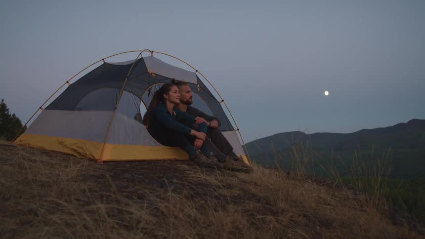 A young married couple sitting close together in a tent looking out and smiling with mountains in the distance