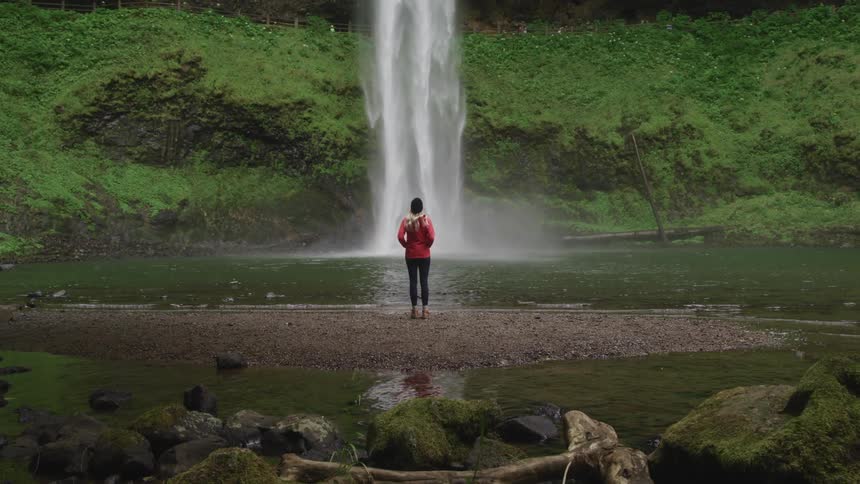 Woman wearing an orange jacket and black hat is watching a waterfall in the forest