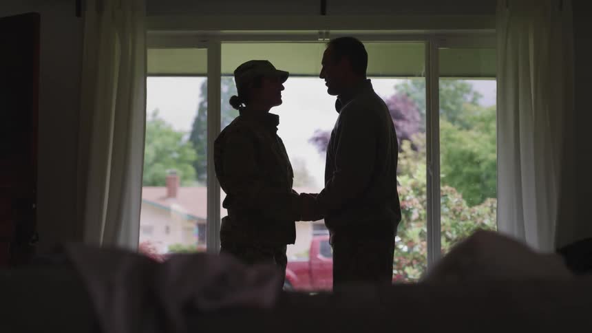 A couple stand in silhoutte holding hands in front of a large window with yard and neighborhood visible