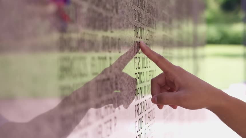 A person runs their finger along a list of names on a memorial outdoors during the day