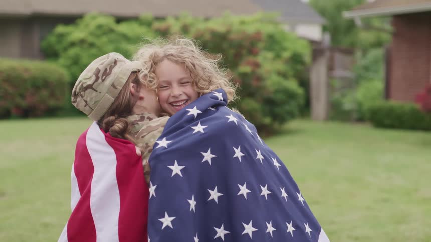 Woman wearing fatigues and girl hug wearing an american flag in front of yard near home