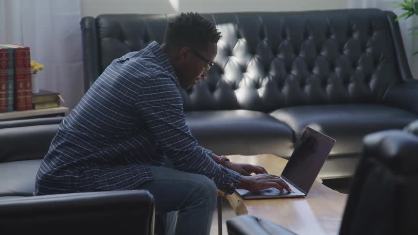 A man in a room sitting on a couch next to a table typing on a laptop
