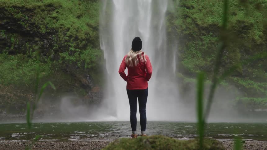 A woman stands and looks out upon a nearby waterfall splashing into a lake below