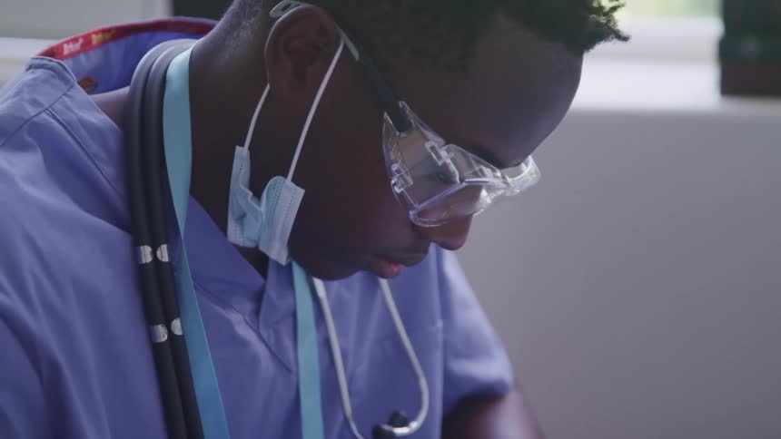 A man wearing blue scrubs with a stethoscope around his neck sitting down inside of a building looking down