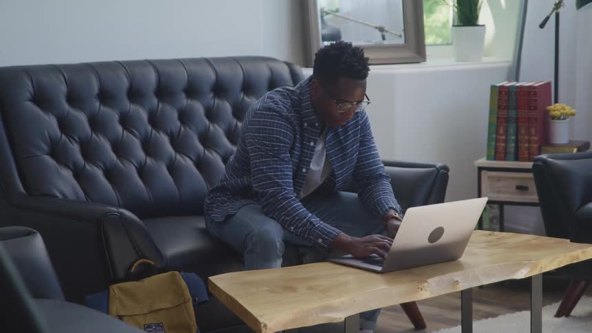 A young man sitting on a black couch working on a laptop in the living room at home